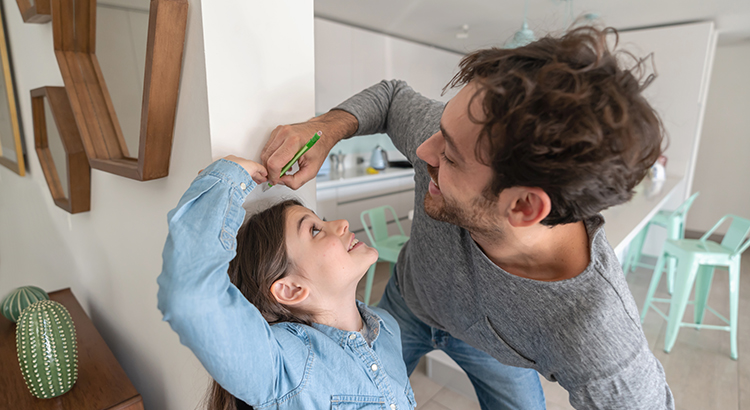 Happy father measuring daughter against the walll while she cheats with her height both smiling