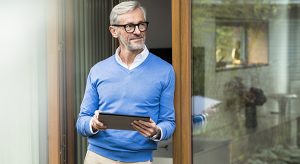 Smiling senior man with grey hair standing in front of his modern design home holding tablet