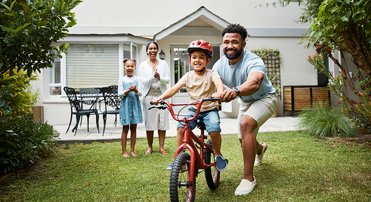 Boy on bicycle learning with proud dad and happy family in their home garden outdoors. Smiling father teaching fun skill, helping and supporting his excited young son to ride, cycle and pedal a bike