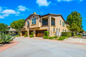 Two-story beige stucco condominium building with attached garages, balconies, and landscaped grounds under a blue sky.