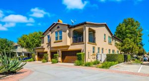 Two-story beige stucco condominium building with attached garages, balconies, and landscaped grounds under a blue sky.